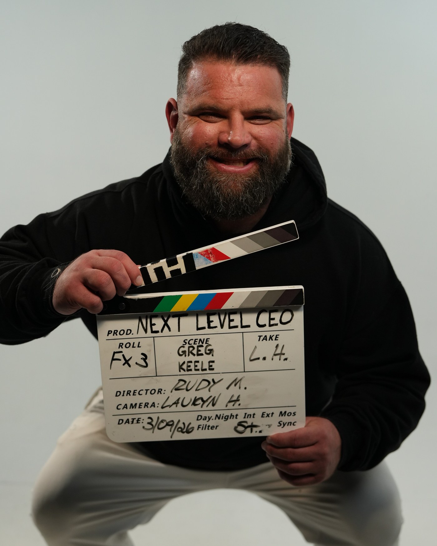 Greg, founder of 208Werx, crouching on a studio backdrop and grinning as he holds up a film slate lettered NEXT LEVEL CEO.